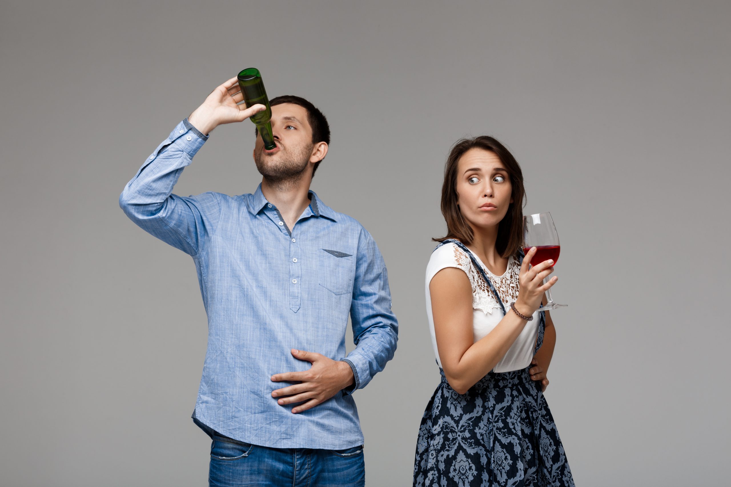 Young beautiful married couple drinking wine and beer over grey background. Copy space.