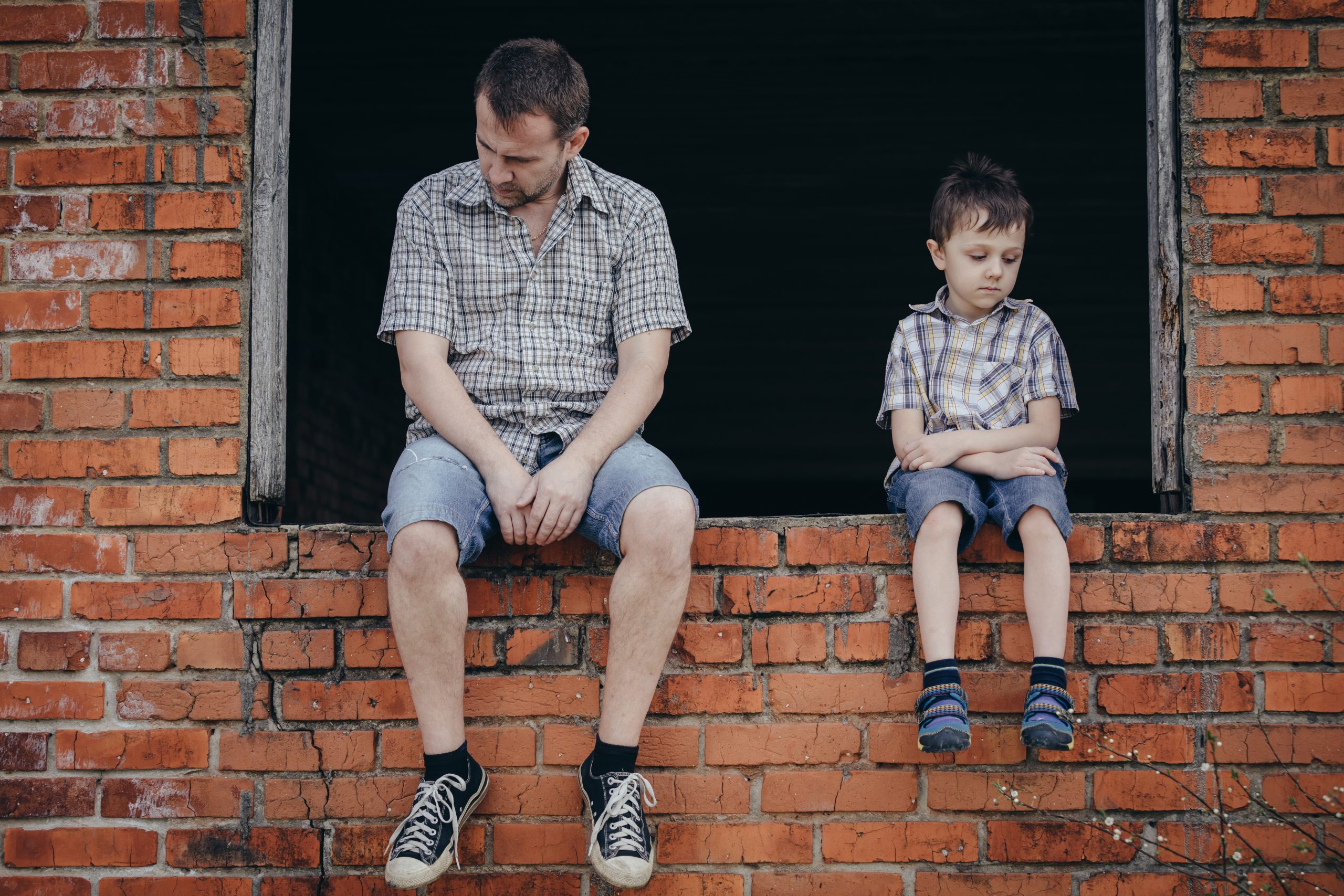 Sad father and son are sitting on the windowsill of the brick building (Emotions) sad man,sad son,loading dock,homeless,windowsill,brick building