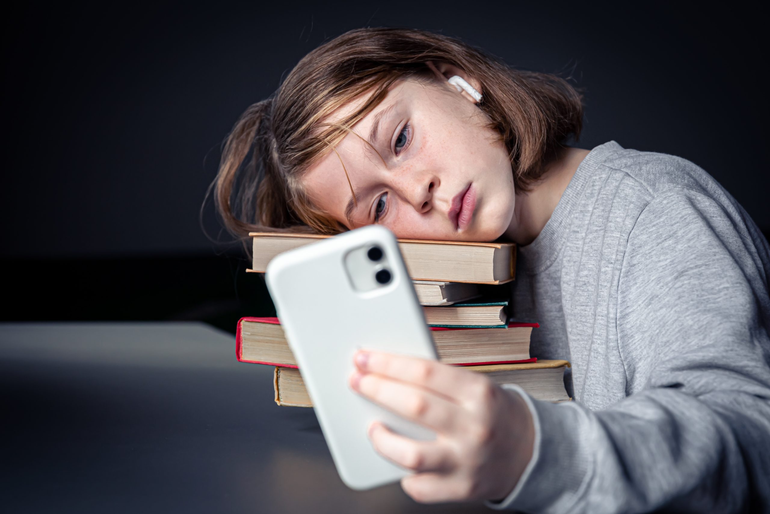 Teenage girl sits with books, but uses a smartphone, boredom from reading, addiction to technology.