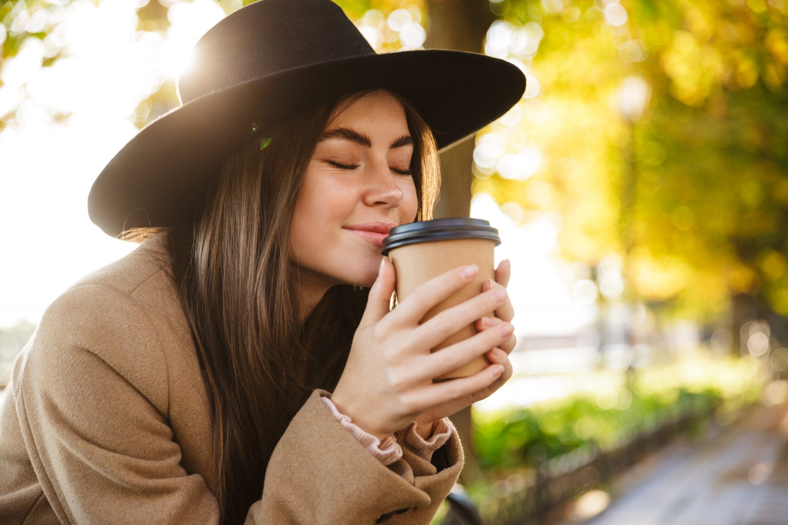 Portrait of young woman wearing coat and hat drinking coffee while walking in autumn park