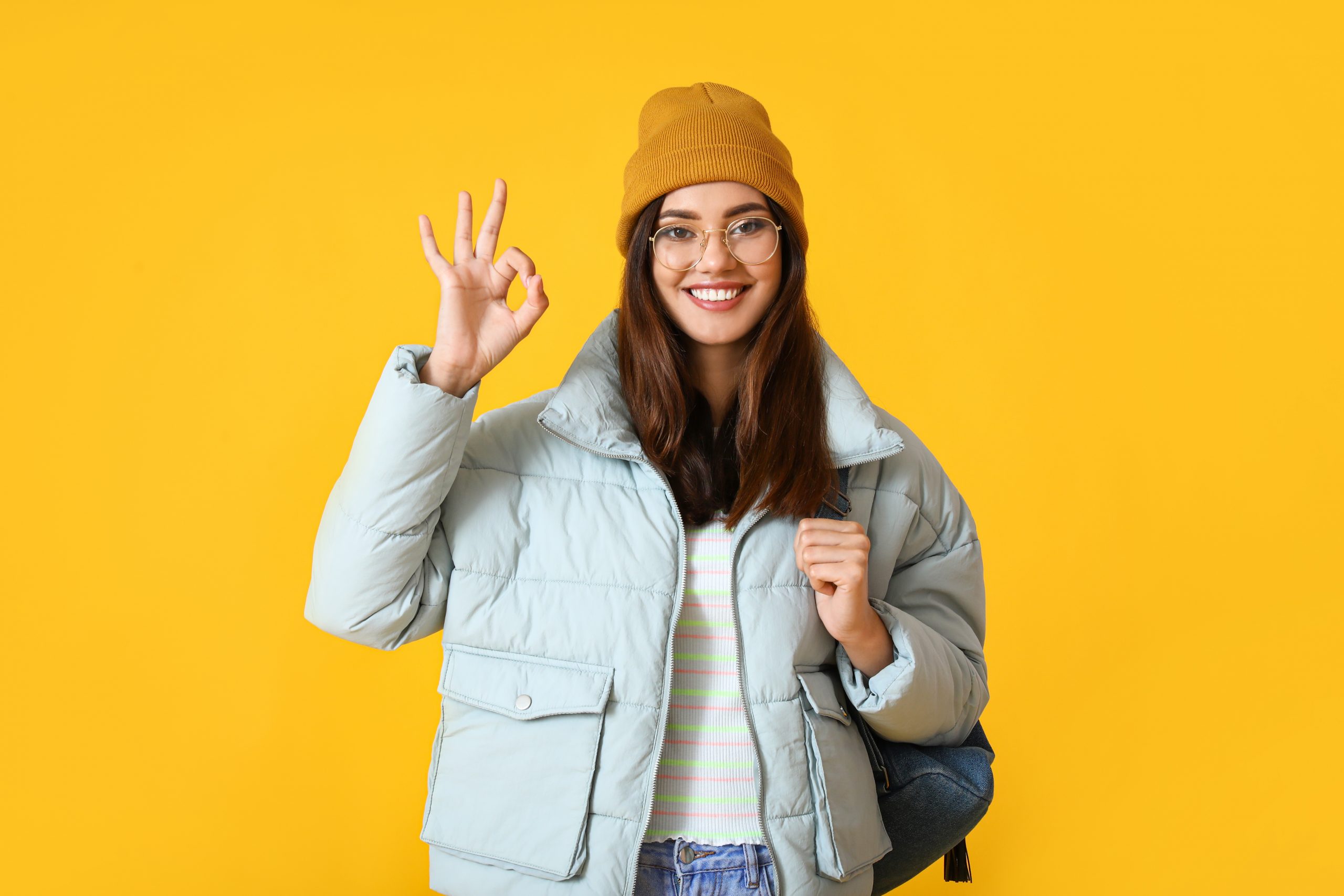 Pretty young woman in warm clothes showing OK on color background