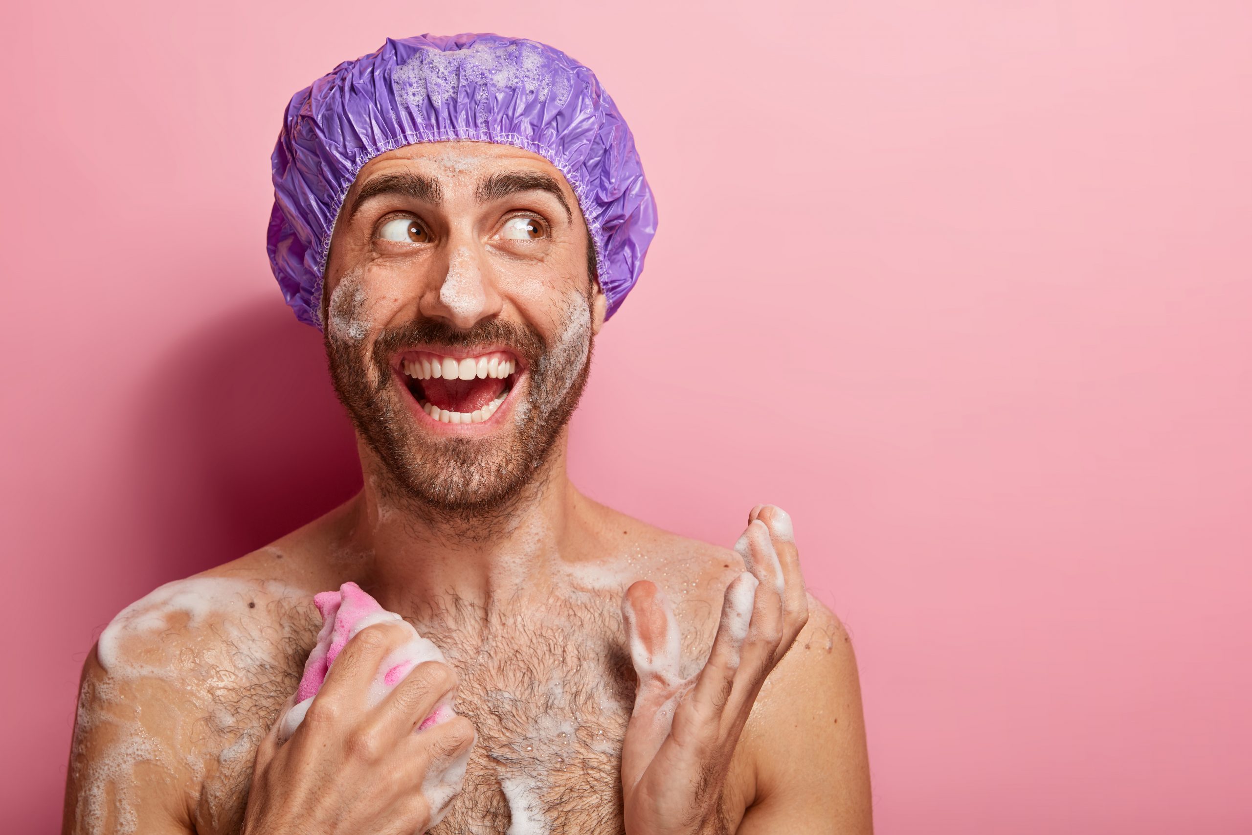 Body care concept. Happy guy takes shower, relaxes after stressed work, has wet skin with foam, rubs chest with sponge, looks joyfully aside, isolated on pink wall, gets energy, washes stress away