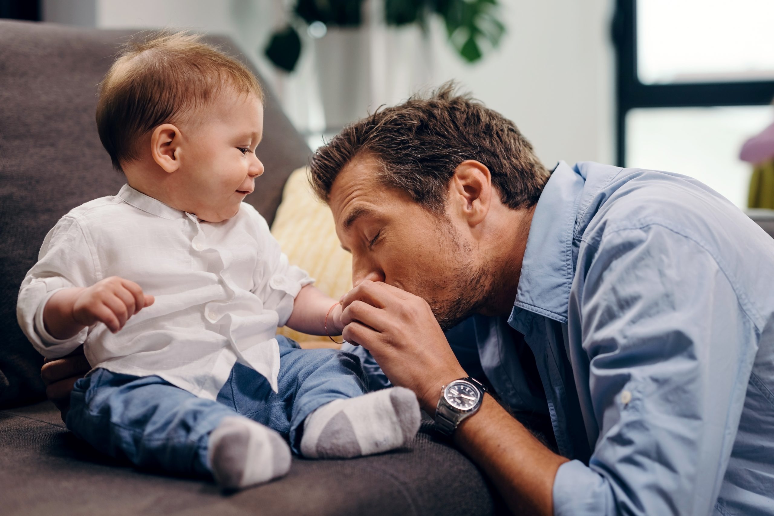 Affectionate father kissing baby son's hand while enjoying in time with him at home.