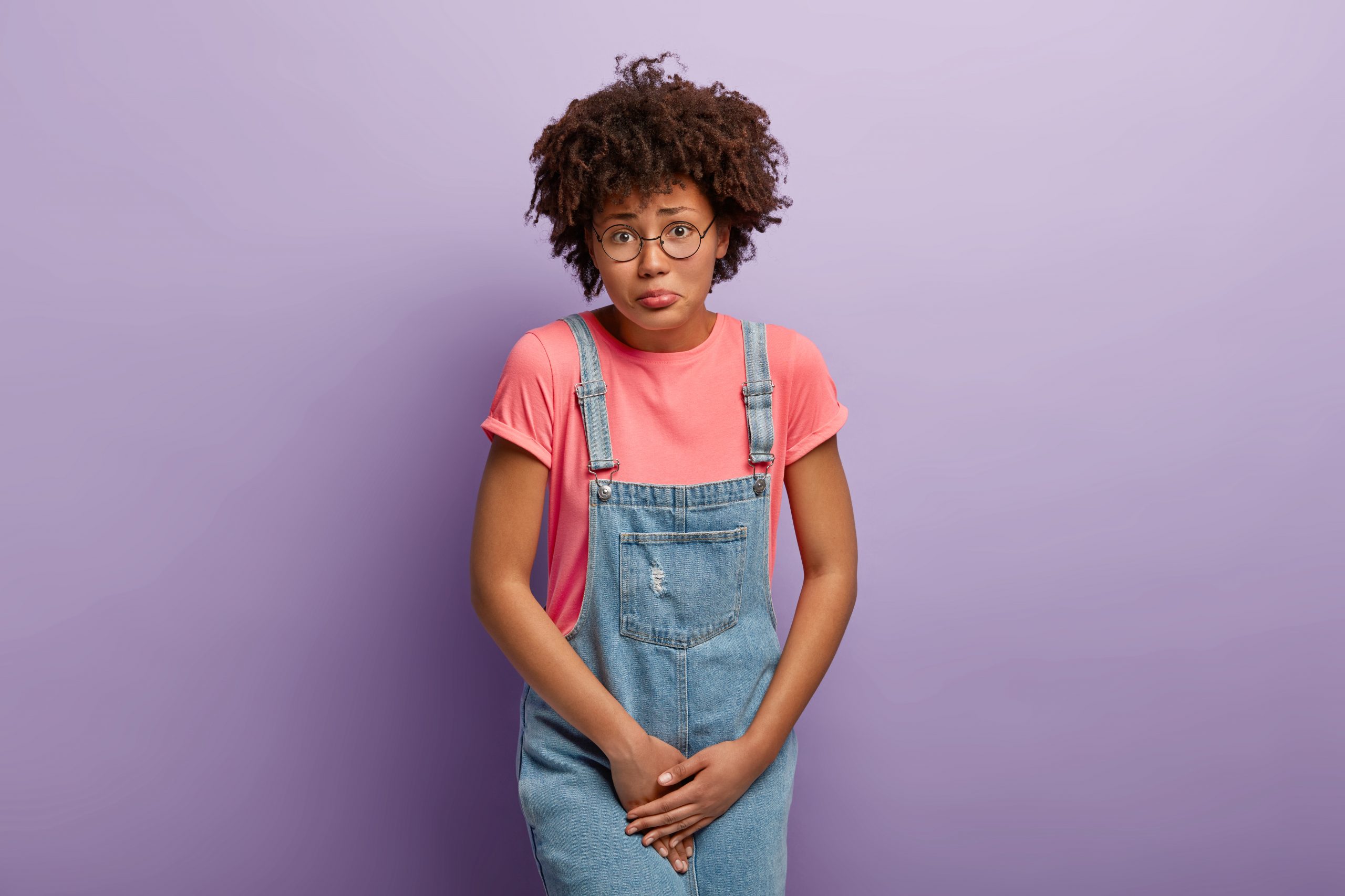 Medical problem, incontience concept. Displeased Afro American woman hods crotch, waits near closed WC, needs toilet, wants to pee, feels discomfort and pain in urinary bladder, wears denim sarafan