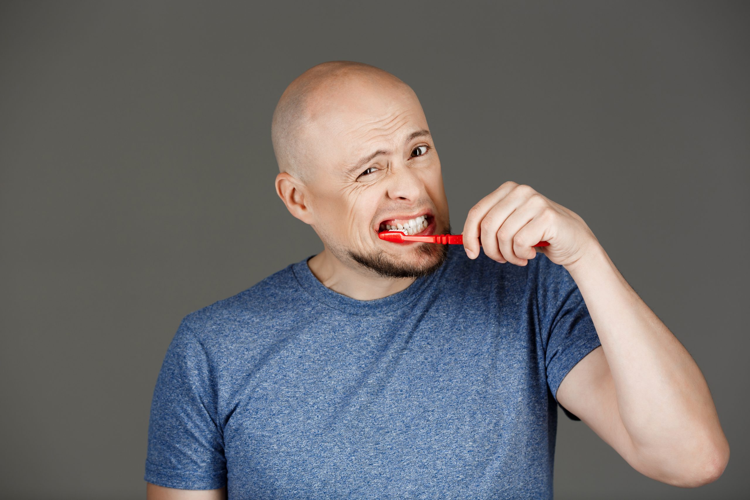 Portrait of funny handsome middle-aged man in grey shirt brushing teeth over dark background. Copy space.