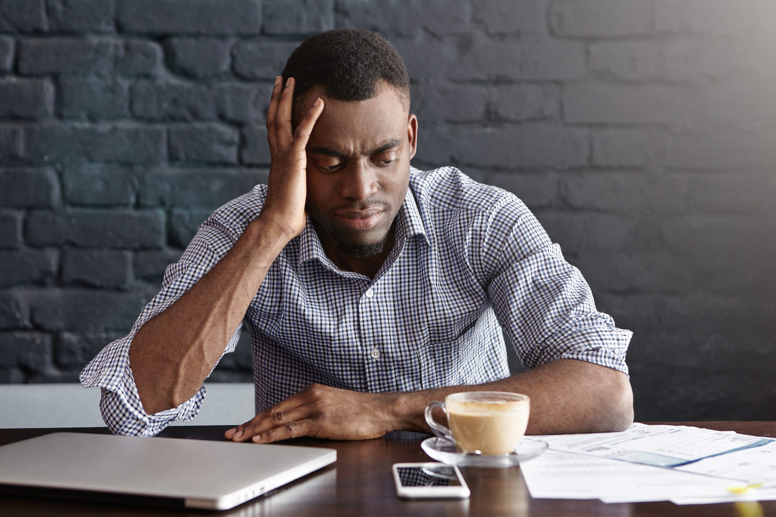 Unhappy African businessman feeling stressed and frustrated, facing financial troubles, leaning elbow on table with mug, laptop, cell phone and papers while doing finances during coffee break