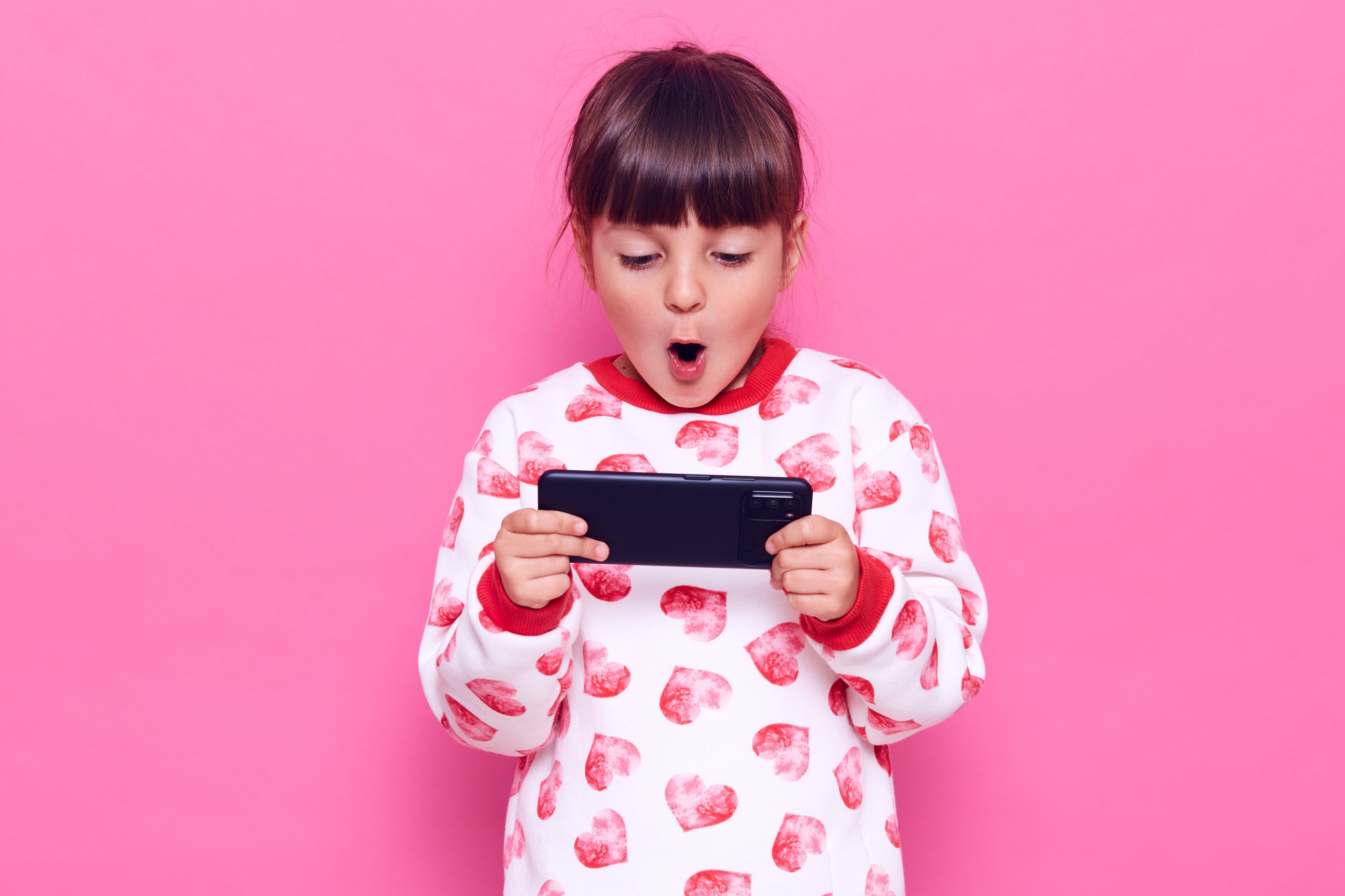 Little dark haired charming girl with dark hair holding smart phone with opened mouth, playing games, being surprised of result, posing isolated over pink background.