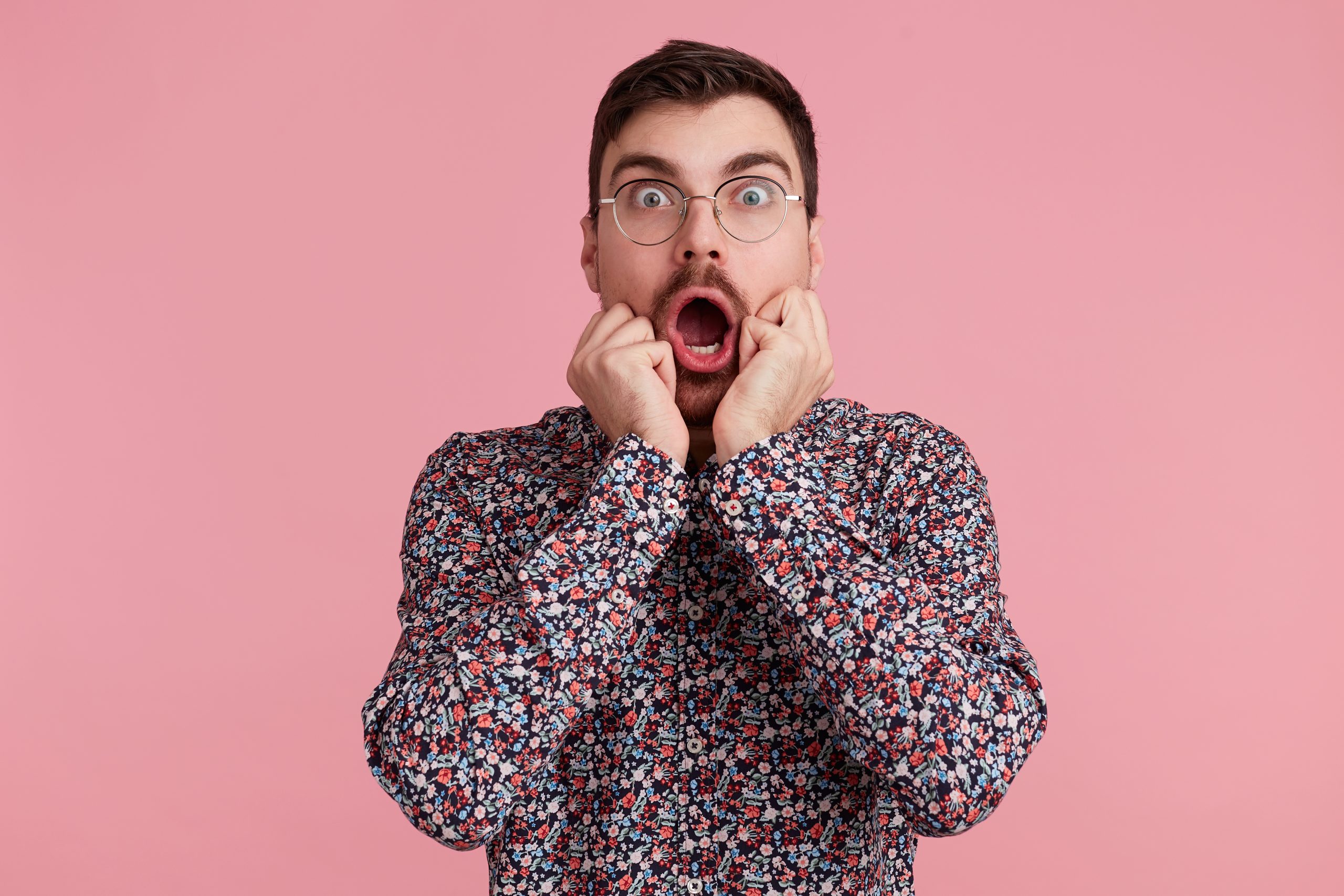 Portrait of young shocked bearded man in glasses, wearing in colorful shirt, with wide open mouth, bites finger nails. Isolated over pink background. People and emotions concept.